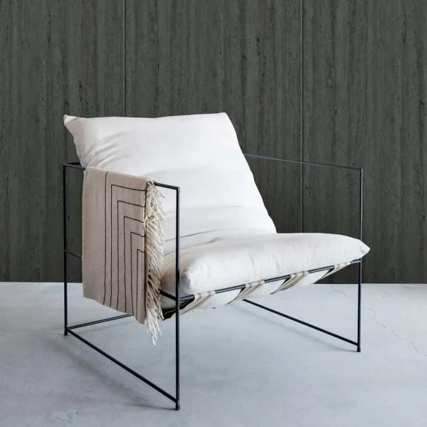 A modern black metal wire-frame lounge chair with off-white cushions and a beige throw blanket draped over the arm, set against a dark textured wall and light concrete floor.