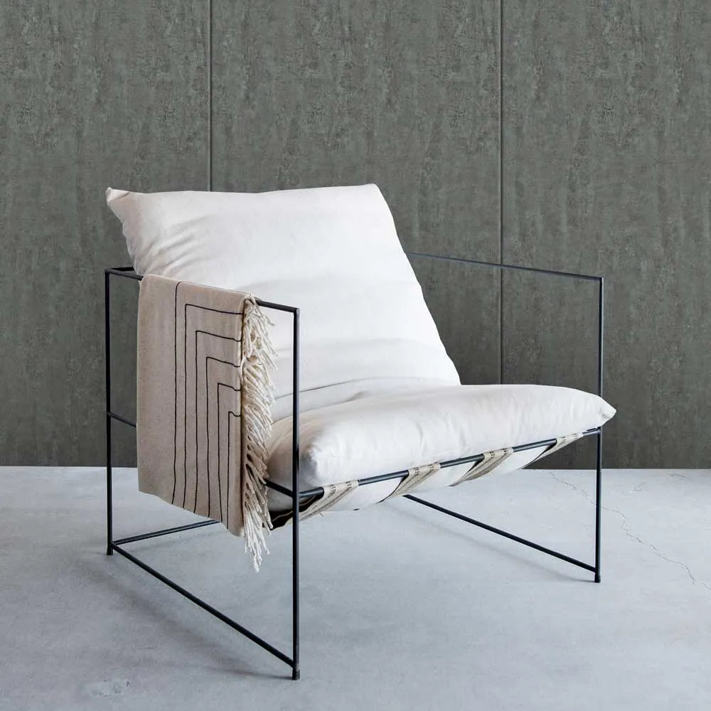 A modern black metal wire-frame lounge chair with thick white cushions, styled with a beige fringed throw blanket, set against a textured gray wall and concrete floor.