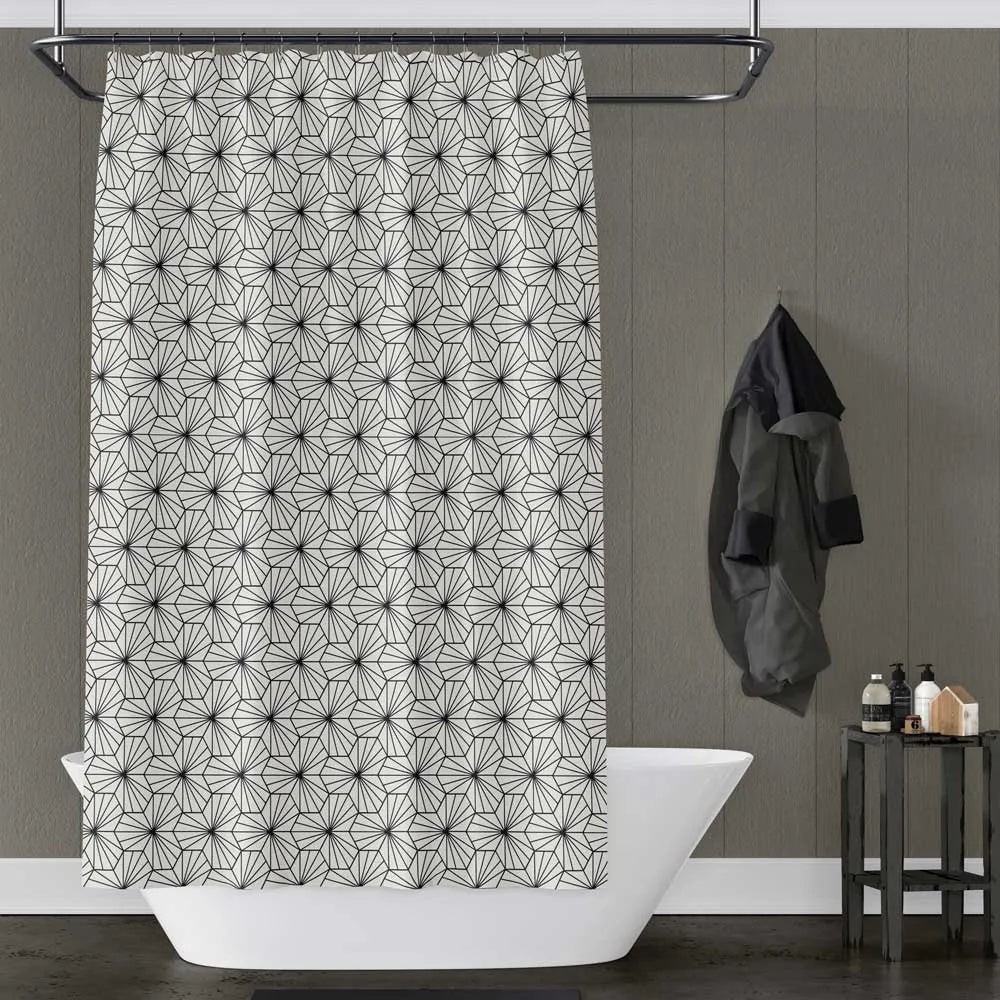 A shower curtain with a repeating black geometric starburst pattern on a white background, hanging in a bathroom setting with a white bathtub, gray paneled wall, and a small side table.