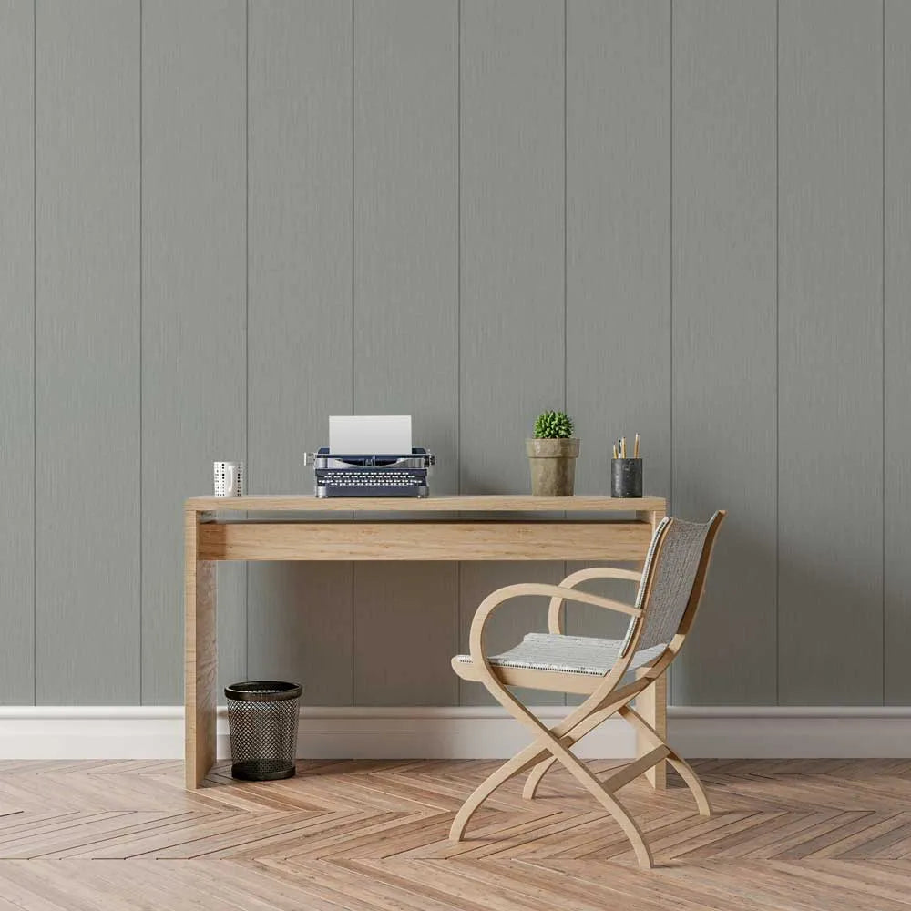 A minimalist home office setup featuring a light oak wood desk and a matching wooden folding chair with a woven seat, set against a gray wood-paneled wall on a herringbone wood floor.