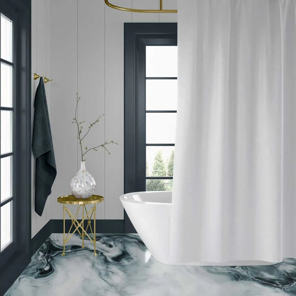A white fabric shower curtain hanging in a modern bathroom with a freestanding white tub, dark gray window frames, a small gold side table with a vase, and a marble-patterned floor.