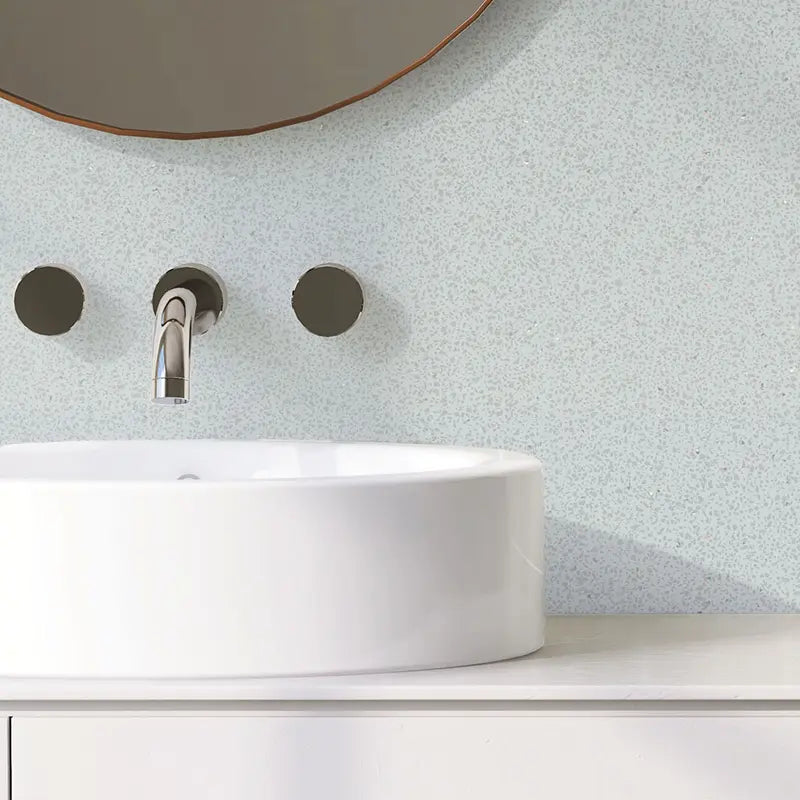 A close-up of a modern bathroom setup featuring a white ceramic vessel sink, a chrome wall-mounted faucet, and two matching wall-mounted controls against a light blue speckled backsplash. A portion of a round mirror with a copper-toned frame is visible at the top.