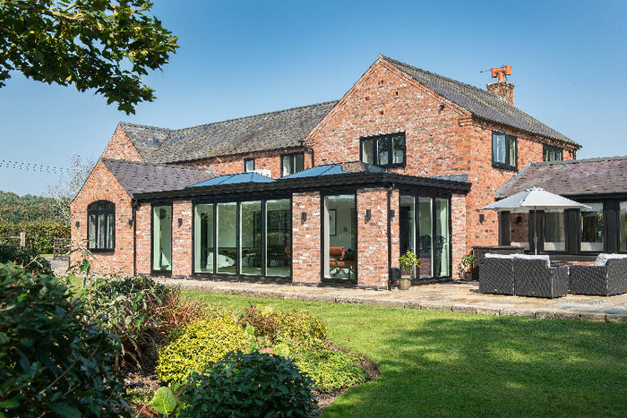 Brick house with large glass extension and outdoor seating area on a sunny day.