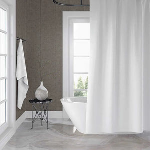 A white fabric shower curtain hanging in a modern bathroom with a freestanding white tub, gray-brown paneled walls, a small black side table with a vase, and a towel on a wall hook.