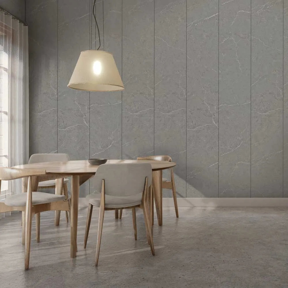 A modern dining room scene with a round wooden table, four upholstered chairs, a hanging pendant lamp, and a gray paneled wall behind them.