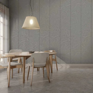 A modern dining room scene with a round wooden table, four upholstered chairs, a hanging pendant lamp, and a gray paneled wall behind them.
