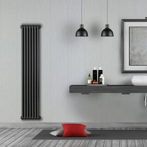 A tall, dark charcoal gray multi-bar radiator mounted on a light gray textured wall in a modern bathroom setting, with a floating shelf, two black pendant lights, and a gray wood floor below.