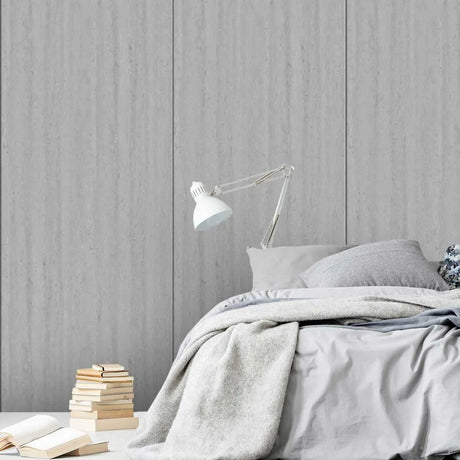 A styled bedroom scene with a bed dressed in light gray bedding, a white adjustable desk lamp, and a stack of books on a bedside surface against a gray wood-paneled wall.