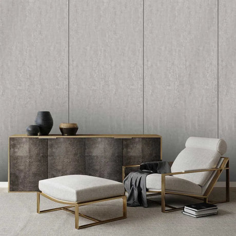 A modern living room scene featuring a light beige upholstered armchair with a gold metal frame, a matching light beige ottoman with a gold frame, and a dark wood sideboard against a textured gray paneled wall.