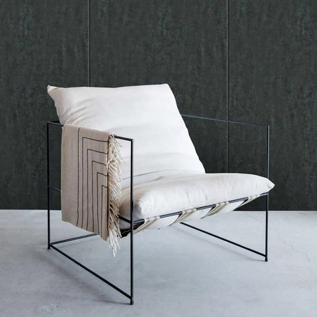 A modern black metal wire-frame lounge chair with thick white cushions and a beige throw blanket draped over the arm, set against a dark textured wall and light concrete floor.