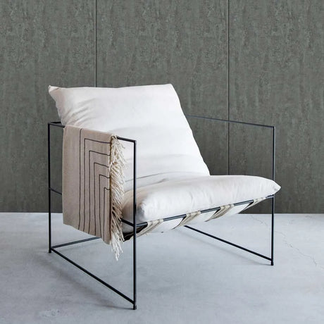 A modern black metal wire-frame lounge chair with thick white cushions, styled with a beige fringed throw blanket, set against a textured gray wall and concrete floor.