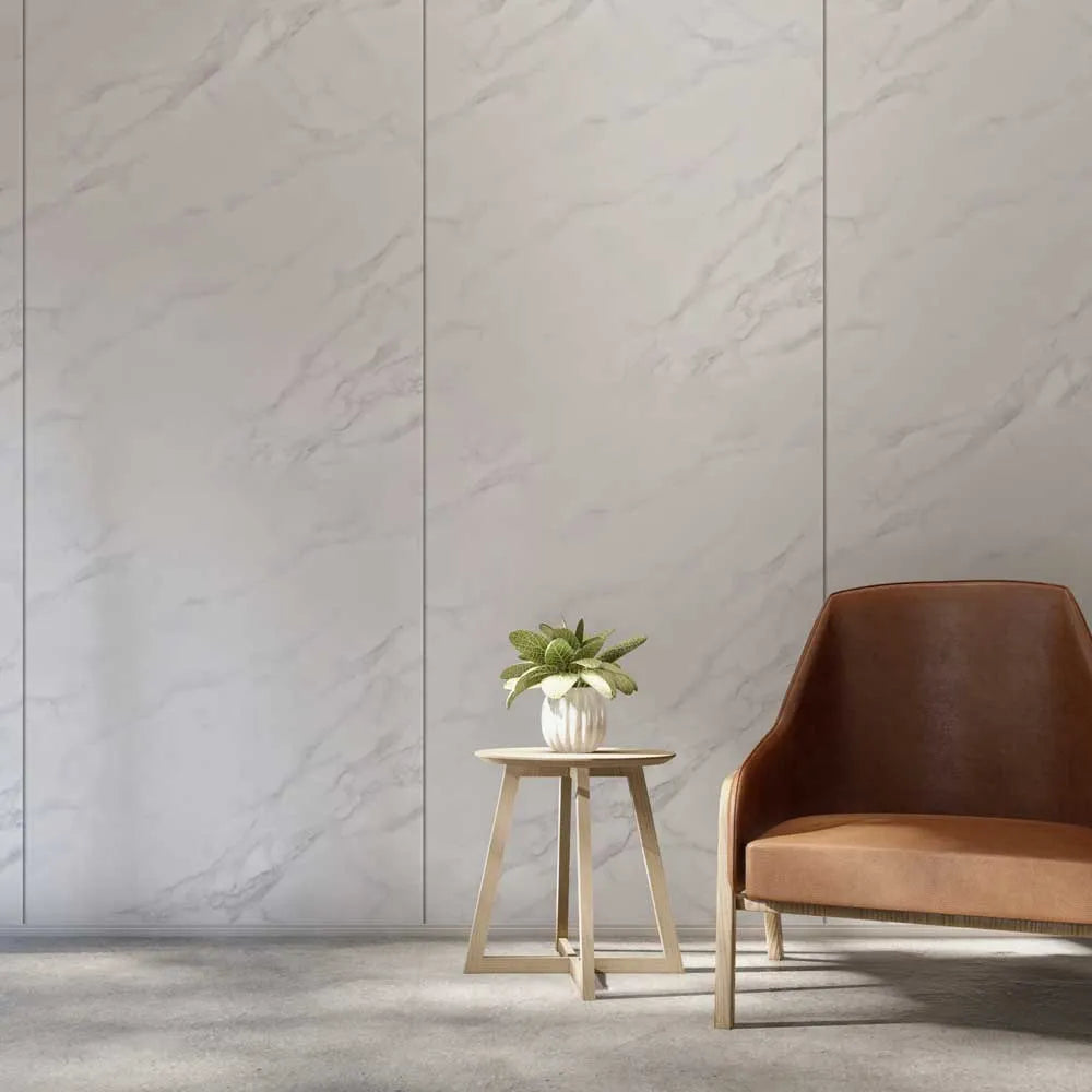A modern interior scene with a brown leather armchair next to a small round wooden side table holding a potted green plant, set against a large light gray marble-patterned wall.
