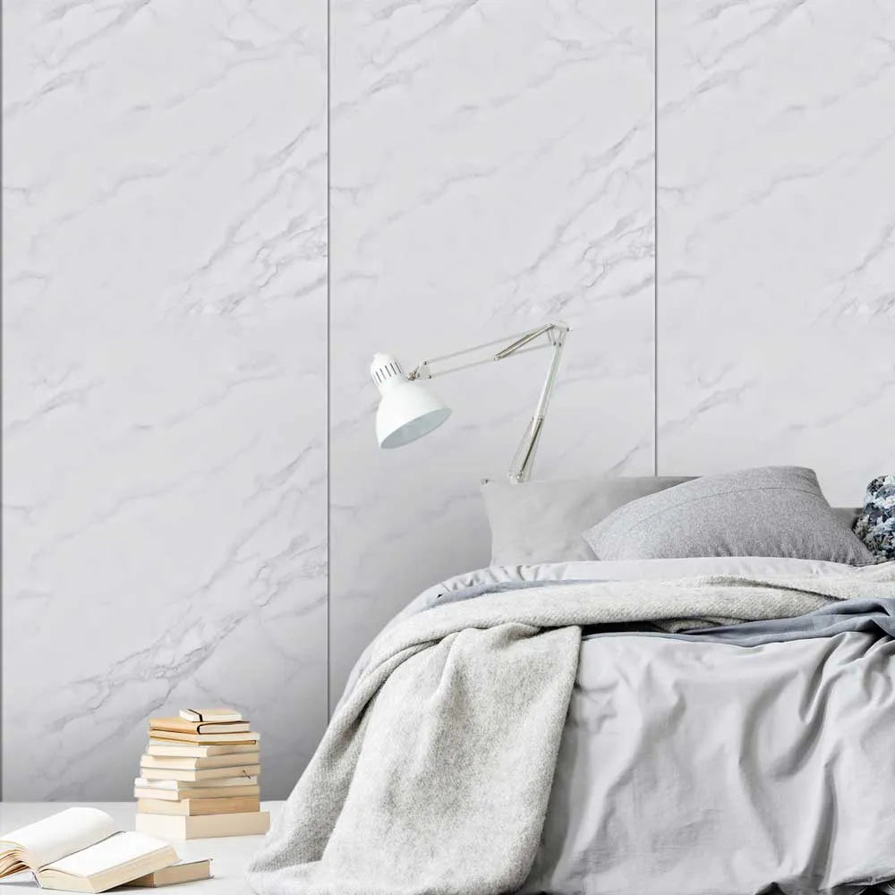 A styled bedroom scene with a bed dressed in light gray bedding, a white adjustable desk lamp, stacked books, and a wall covered in large white marble-look panels.