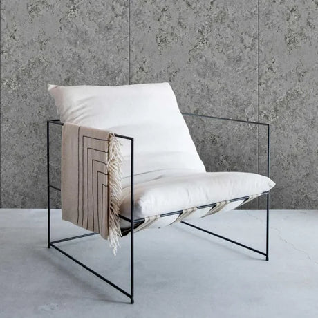 A modern black metal frame lounge chair with white cushions and a beige fringed throw blanket, set against a textured gray wall and concrete floor.