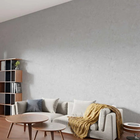A modern living room scene featuring a large, textured light gray wall, a light gray sofa with white and blue pillows, a mustard yellow throw blanket, and a wooden bookshelf on the left.
