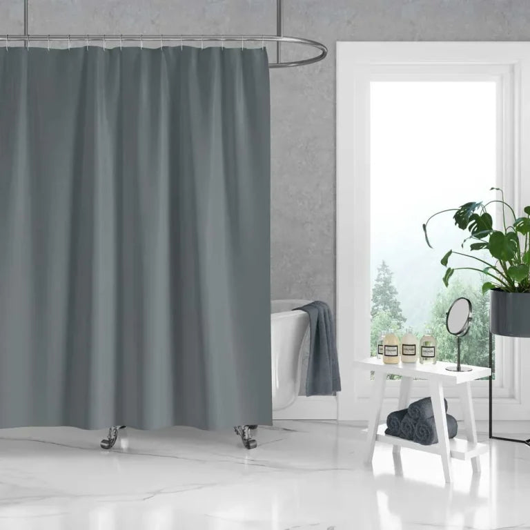 A gray fabric shower curtain hanging in a bright, modern bathroom with a white tub, small white stool, window view, and potted plant.