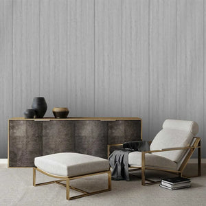 A modern living room scene featuring a light beige upholstered armchair with a gold metal frame, a matching light beige ottoman with a gold frame, and a dark wood sideboard against a textured gray paneled wall.