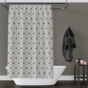 A shower curtain with a repeating black geometric starburst pattern on a white background, hanging in a bathroom setting with a white bathtub, gray paneled wall, and a small side table.