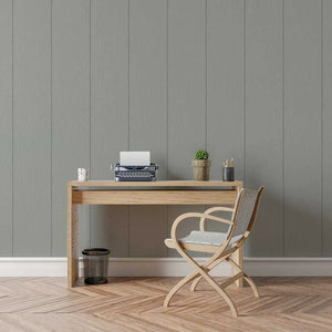 A minimalist home office setup featuring a light oak wood desk and a matching wooden folding chair with a woven seat, set against a gray wood-paneled wall on a herringbone wood floor.