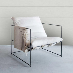 A modern black metal wire-frame lounge chair with white cushions, styled with a beige throw blanket featuring black geometric stripes and fringe, set against a light gray tiled wall and concrete floor.