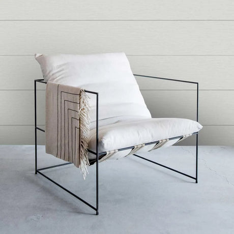 A modern black metal wire-frame lounge chair with white cushions, styled with a beige throw blanket featuring black geometric stripes and fringe, set against a light gray tiled wall and concrete floor.