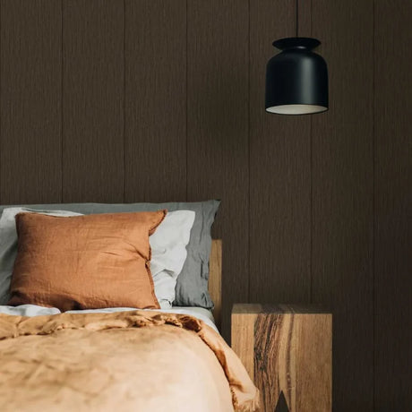 A modern bedroom scene featuring a bed with a rust-colored linen pillow and beige bedding, set against a dark brown wood-paneled wall under a black pendant light.
