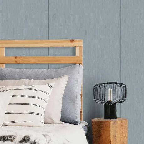 A styled bedroom corner with a light wood bed frame, gray and striped pillows, a black wire lamp, and a blue-gray paneled wall.