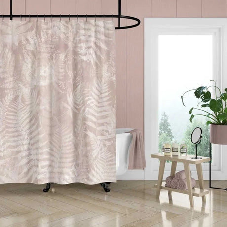 A bathroom scene showing a blush pink shower curtain with a light beige fern leaf pattern, hanging in a room with light wood floors, a white clawfoot tub, and a small wooden side table.