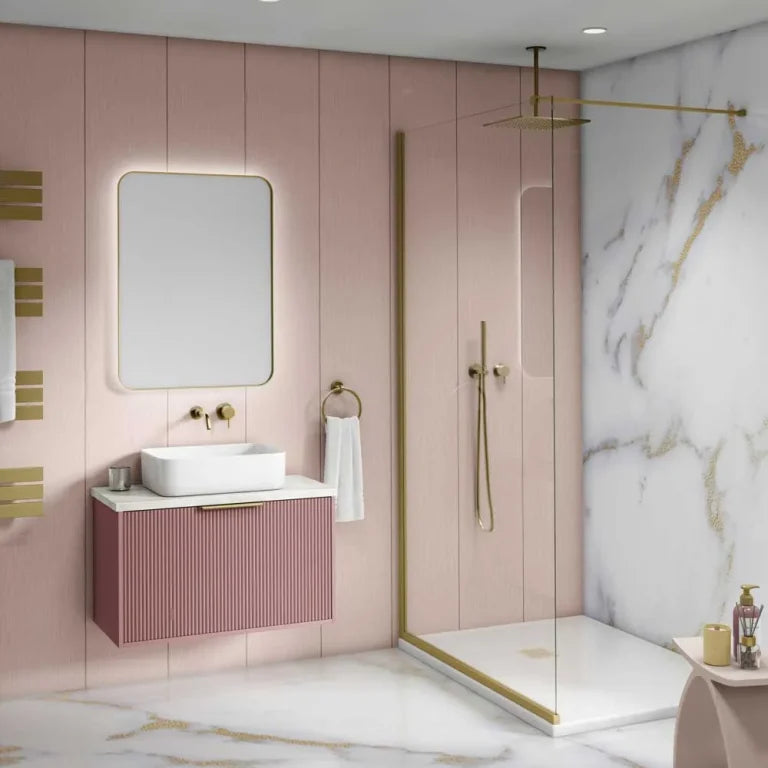 A modern bathroom interior featuring a floating pink vanity with a white vessel sink, a large rounded rectangular mirror, and a pink ribbed cabinet with gold hardware. A glass shower door, gold shower fixtures, and a white marble wall with gold veining are visible in the background.