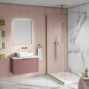A modern bathroom interior featuring a floating pink vanity with a white vessel sink, a large rounded rectangular mirror, and a pink ribbed cabinet with gold hardware. A glass shower door, gold shower fixtures, and a white marble wall with gold veining are visible in the background.