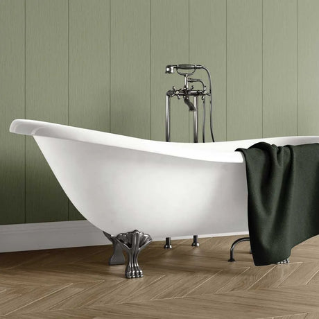 A freestanding white clawfoot bathtub with ornate silver metal feet and a matching chrome faucet set, shown in a bathroom with olive green paneled walls and wood herringbone flooring. A dark green towel is draped over the side of the tub.