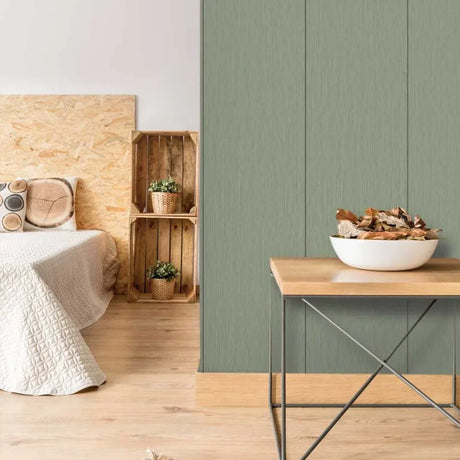 A styled interior scene showing a sage green vertical wood-look wall paneling installed on a wall in a bedroom/living space, alongside a light wood bed, wooden crates, a side table, and a bowl of dried leaves.