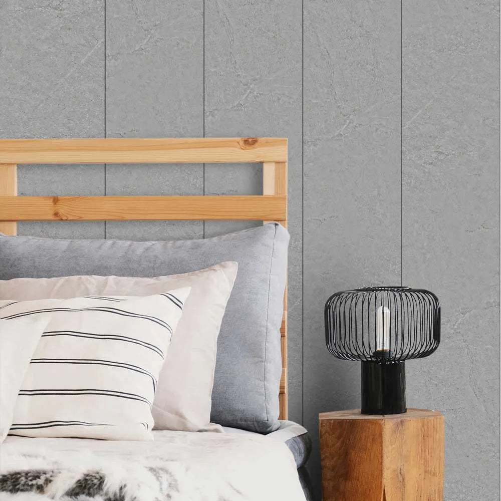 A styled bedroom scene showing a bed with light wood bedding, gray pillows, and a black wire-frame table lamp on a wooden side table against a gray paneled wall.