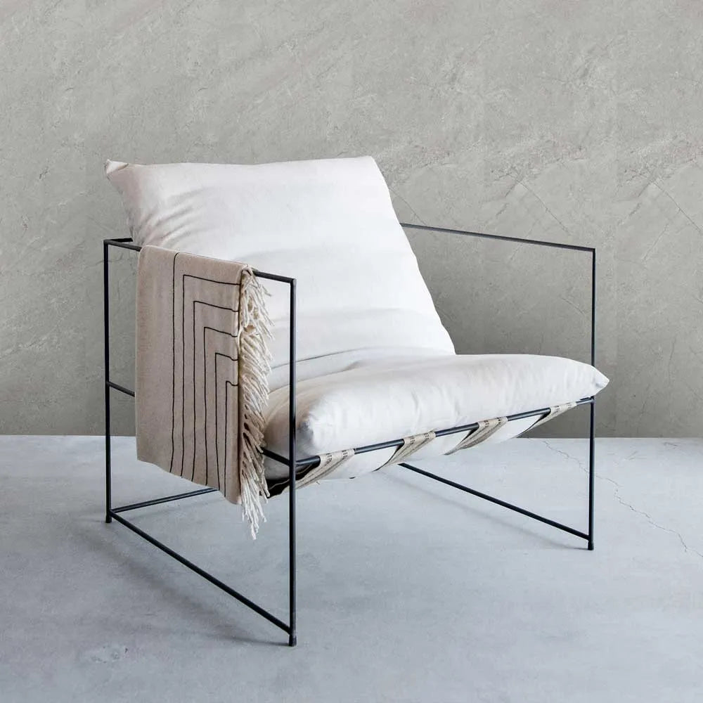 A modern black metal wire chair with white cushions and a beige fringed throw blanket featuring black geometric stripes, set against a textured gray concrete wall and floor.