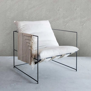 A modern black metal wire chair with white cushions and a beige fringed throw blanket featuring black geometric stripes, set against a textured gray concrete wall and floor.
