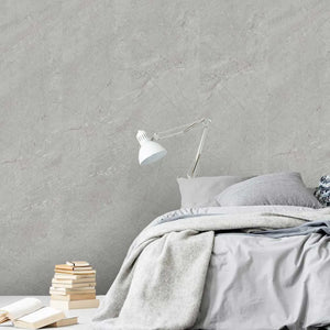 A styled bedroom scene with a bed dressed in light gray bedding, a white adjustable desk lamp, stacked books, and an open book on a white surface against a textured light gray wall.