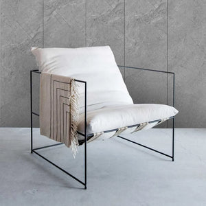 A modern black metal wire armchair with white cushions and a beige throw blanket draped over the side, set against a gray stone-textured wall and floor.