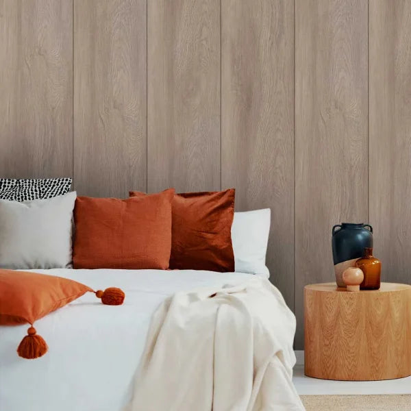 A styled bedroom scene with a bed dressed in white bedding and multiple pillows, including rust-orange and white accent pillows, set against a light wood paneled wall. A round wooden side table beside the bed holds dark and amber glass vases.