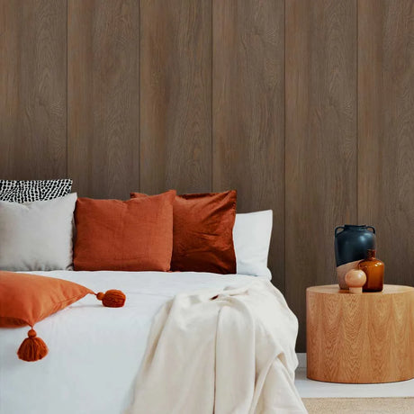 A styled bedroom scene with a bed dressed in white bedding and multiple pillows, including rust-orange and terracotta cushions, set against a brown wood-paneled wall. A round wooden side table holds decorative vases on the right.