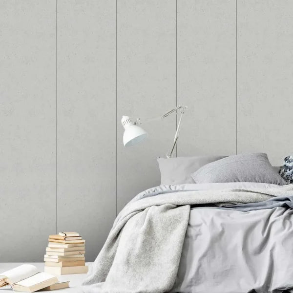 A styled bedroom scene with a bed dressed in light gray bedding, a white adjustable desk lamp, stacked books, and a textured light gray paneled wall behind.