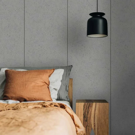 A modern bedroom scene featuring a black pendant light hanging above a bed with a rust-orange linen pillow, white and gray pillows, and a textured beige duvet. The background consists of gray wood-paneled walls, and a wooden nightstand is visible on the right.