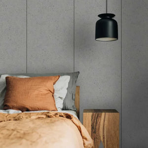 A modern bedroom scene featuring a black pendant light hanging above a bed with a rust-orange linen pillow, white and gray pillows, and a textured beige duvet. The background consists of gray wood-paneled walls, and a wooden nightstand is visible on the right.