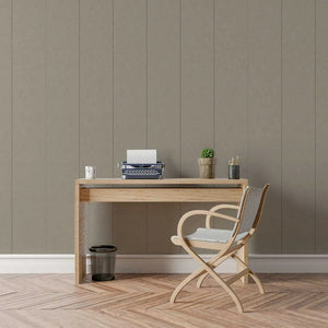 A styled interior scene showing a light wood desk against a taupe paneled wall, with a wooden folding chair, a small potted plant, a typewriter, and a small cup on top. The floor is herringbone wood.