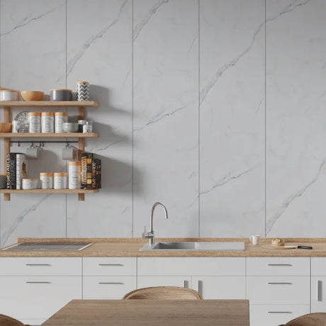 A modern kitchen interior with white marble-look wall panels, white cabinets, a light wood countertop, a built-in sink, and floating wooden shelves holding jars and bowls.