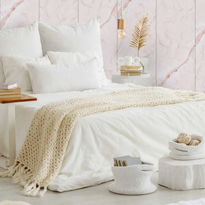 A styled bedroom scene with a bed dressed in white bedding and a cream crocheted throw blanket, set against pink marble wall panels with bedside tables, books, a lamp, and decorative baskets.
