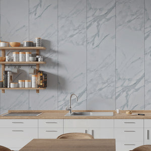A modern kitchen interior with white cabinets, a light wood countertop, a built-in sink, and a large marble-look wall panel behind it. Floating wooden shelves on the left hold jars, bowls, and books.