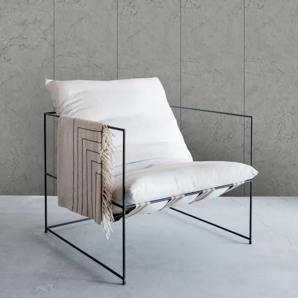 A modern black metal wire-frame lounge chair with white cushions and a beige throw blanket draped over the side, set against a textured gray concrete wall and floor.