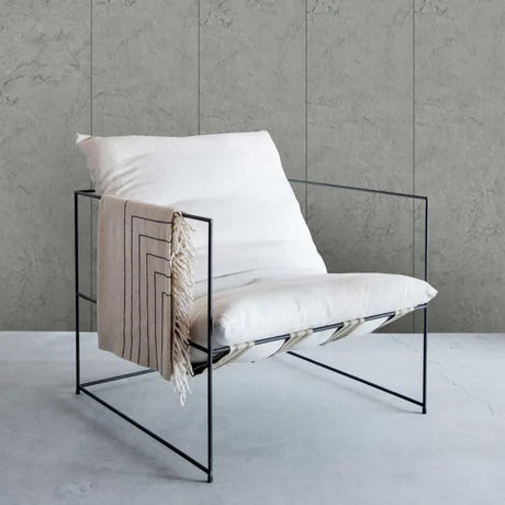A modern black metal wire-frame lounge chair with white cushions and a beige throw blanket draped over the side, set against a textured gray concrete wall and floor.