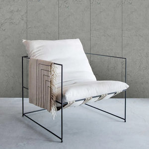 A modern black metal wire-frame lounge chair with white cushions and a beige throw blanket draped over the side, set against a textured gray concrete wall and floor.
