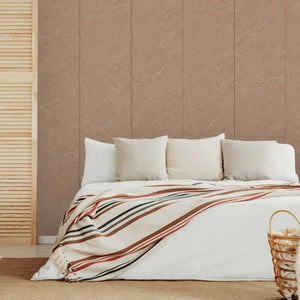 A styled bedroom scene with a bed dressed in white bedding and a cream throw blanket featuring rust, brown, and black stripes. The background features a large wall panel in a warm taupe-brown marble texture, with a light wood slatted screen on the left and a woven basket on the floor in the foreground.